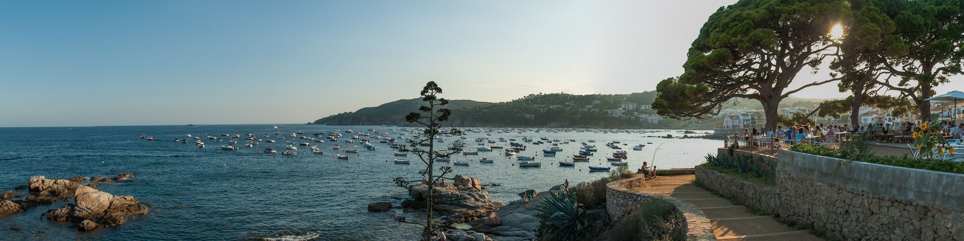 A coastal scene with a stone pathway, rocky shoreline, and numerous boats in the water.