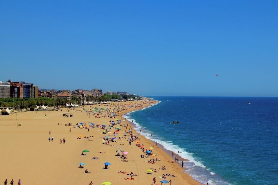 Uma praia cheia de gente, com pessoas se banhando ao sol e nadando, céu azul claro e edifícios ao fundo.