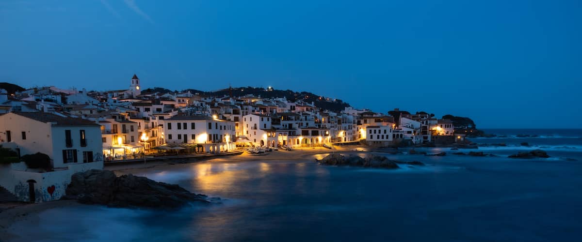 A coastal town at dusk with illuminated buildings, a rocky shoreline, and a calm sea.