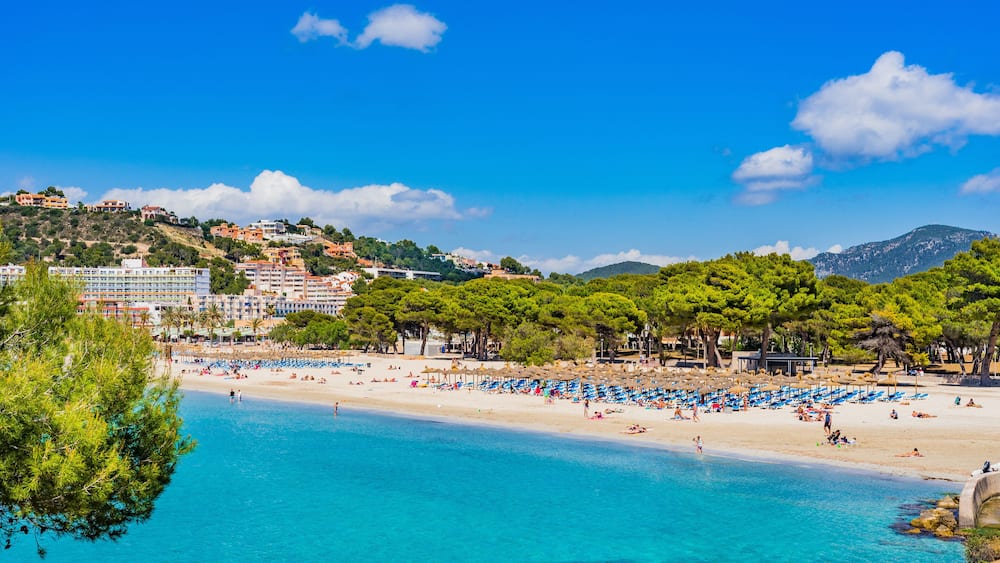 A coastal beach with clear blue water, a sandy shore, and a backdrop of buildings and trees on a hillside.