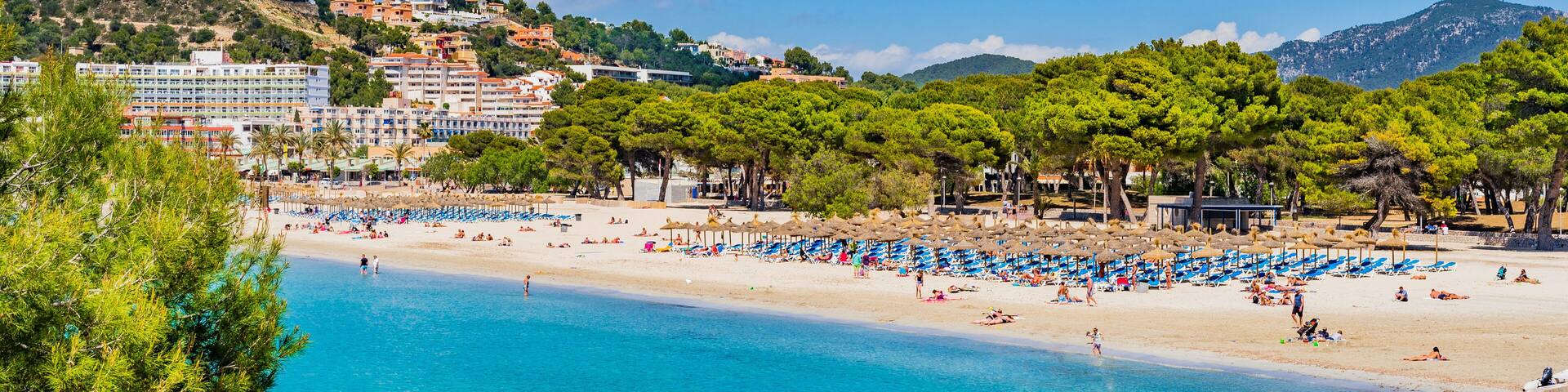 A coastal beach with clear blue water, a sandy shore, and a backdrop of buildings and trees on a hillside.
