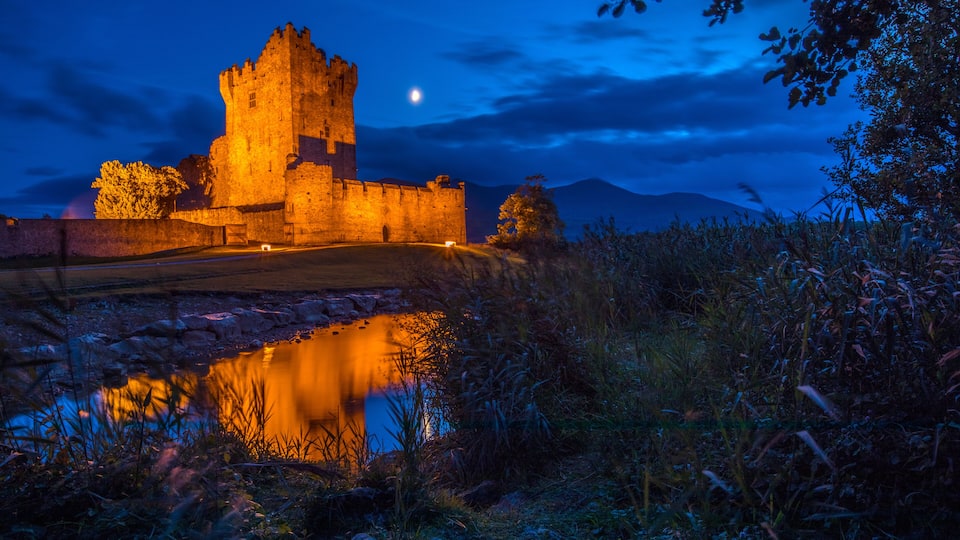 A historic castle at twilight with a moat and a reflective water surface.