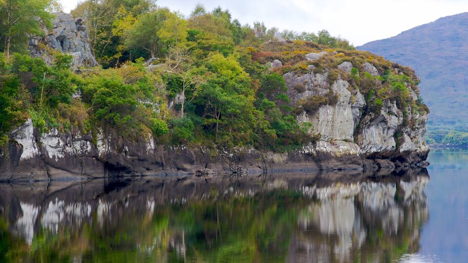 Un acantilado rocoso con abundante vegetación verde y un lago tranquilo que refleja el paisaje.