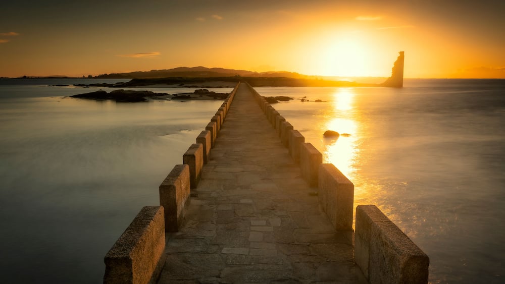 A stone pier leading to a lighthouse by the sea at sunset.