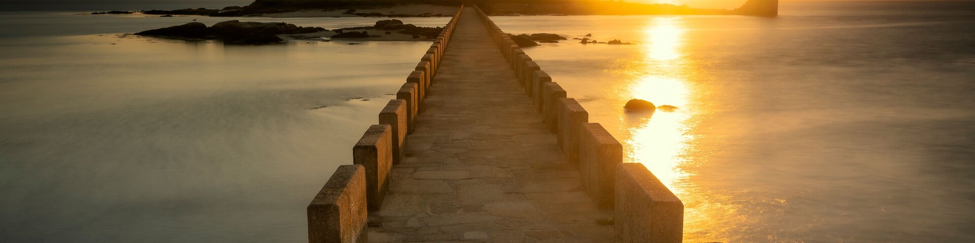 Um cais de pedra que leva a um farol à beira-mar ao pôr do sol.