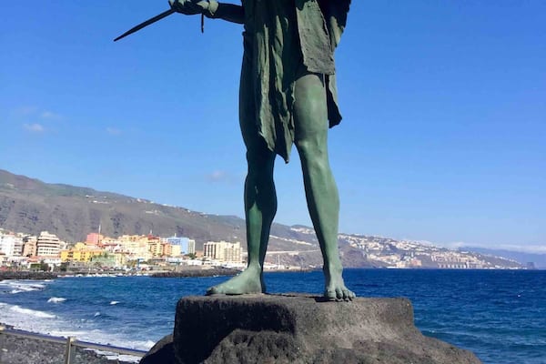 Estátua de um homem segurando uma lança, em cima de uma rocha à beira-mar, com um horizonte urbano ao fundo.