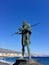 A statue of a man holding a spear, standing on a rock by the sea with a cityscape in the background.