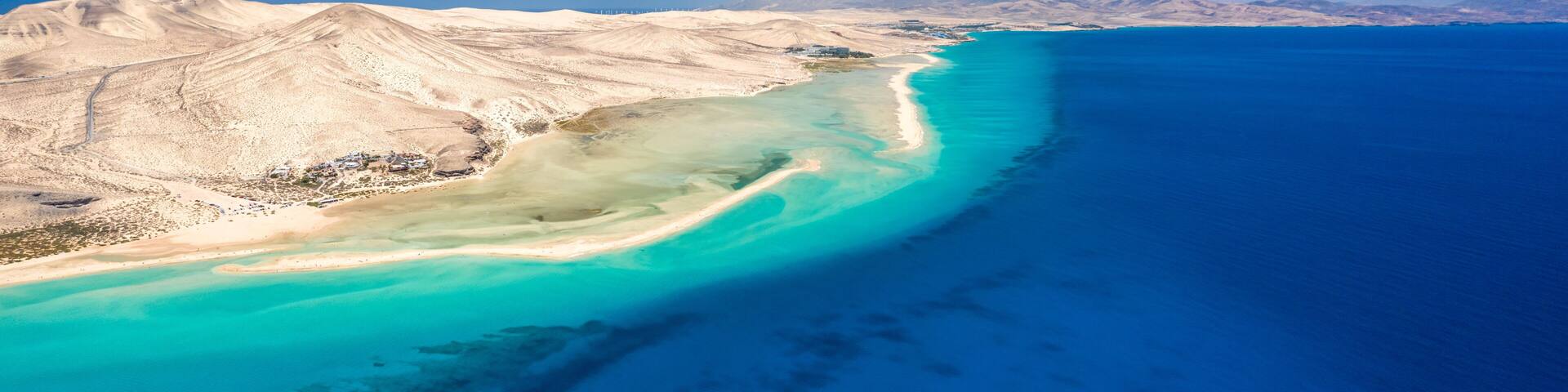 A coastal landscape with a sandy beach, turquoise waters, and distant mountains under a clear blue sky.