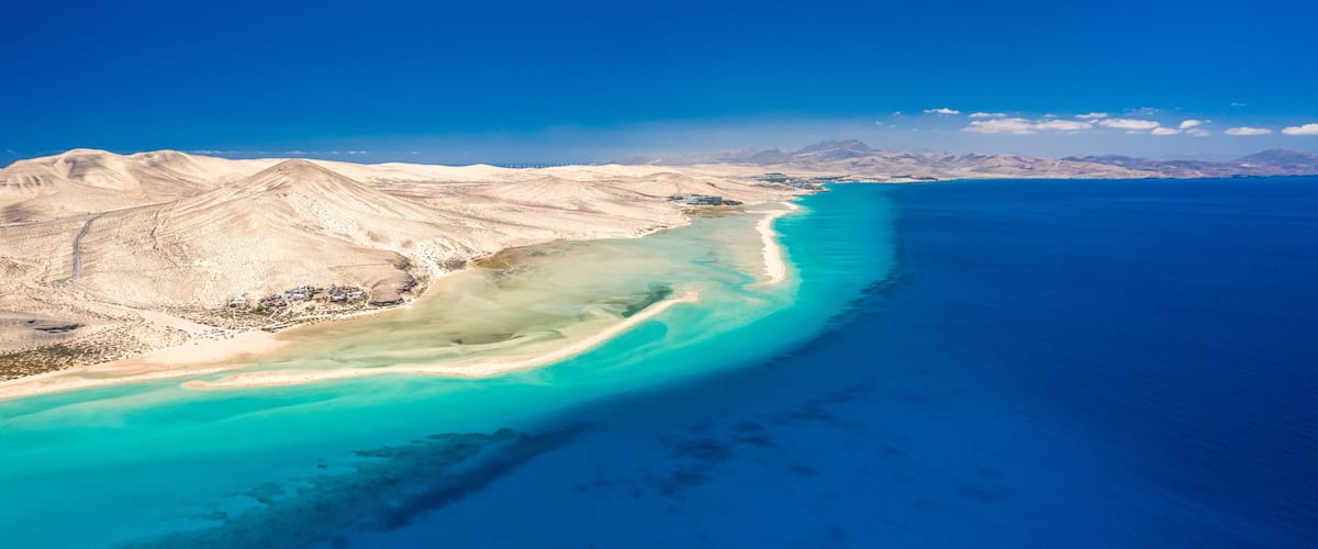 A coastal landscape with a sandy beach, turquoise waters, and distant mountains under a clear blue sky.