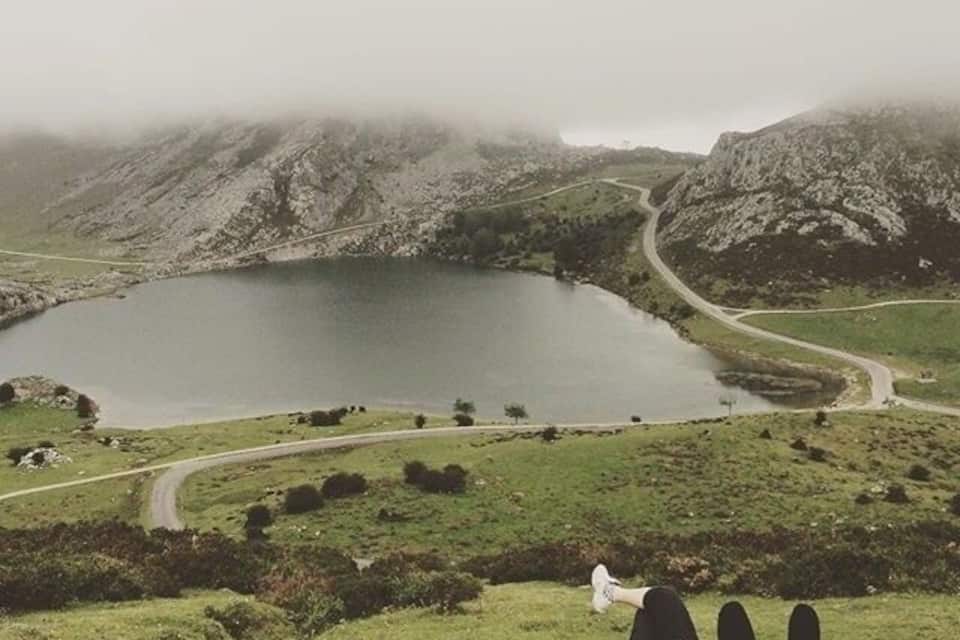 Un lago rodeado de colinas, un camino sinuoso y una persona acostada sobre la hierba.