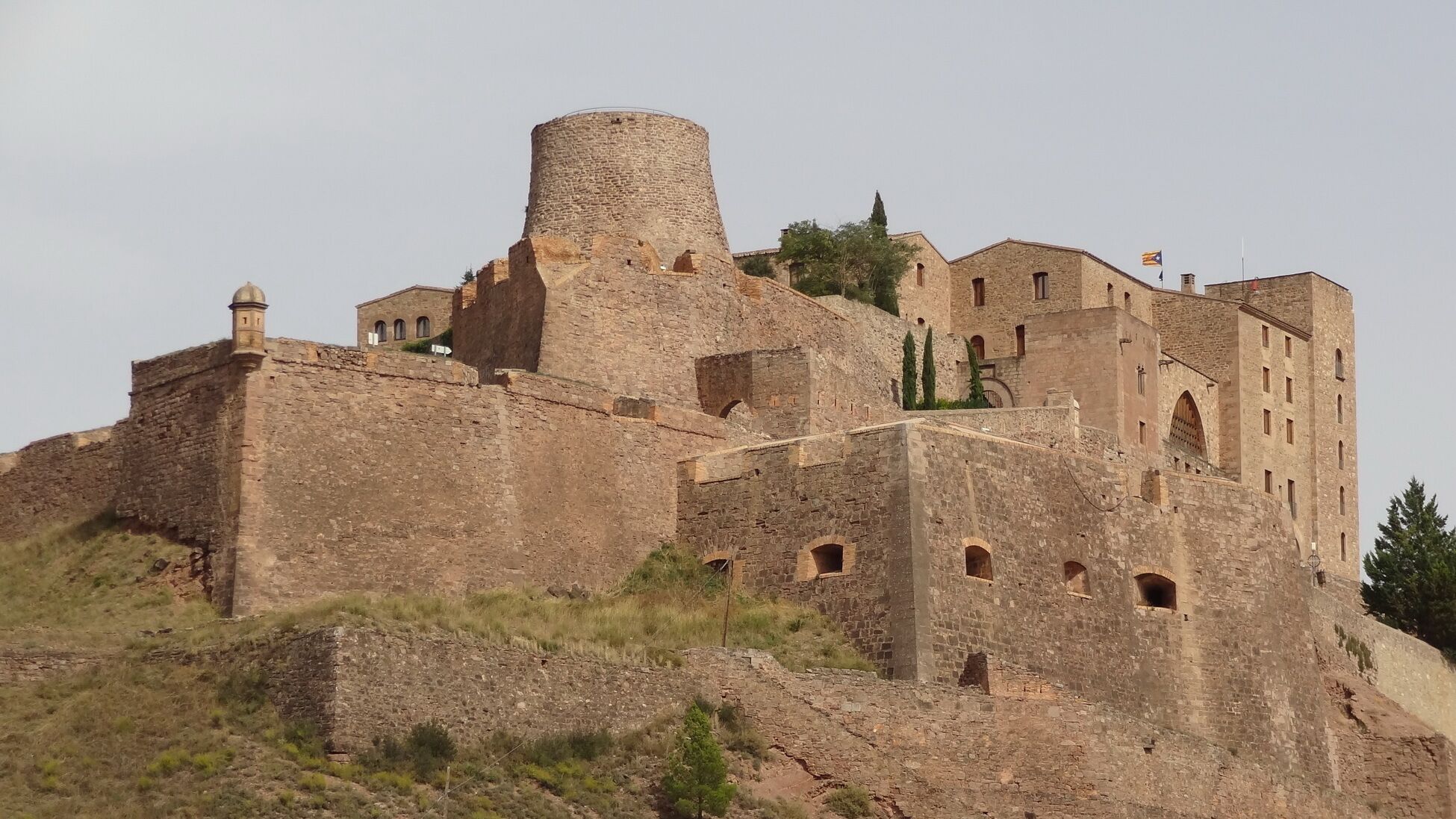 Um castelo de pedra histórico com uma torre redonda imponente, janelas em arco e uma bandeira no topo.