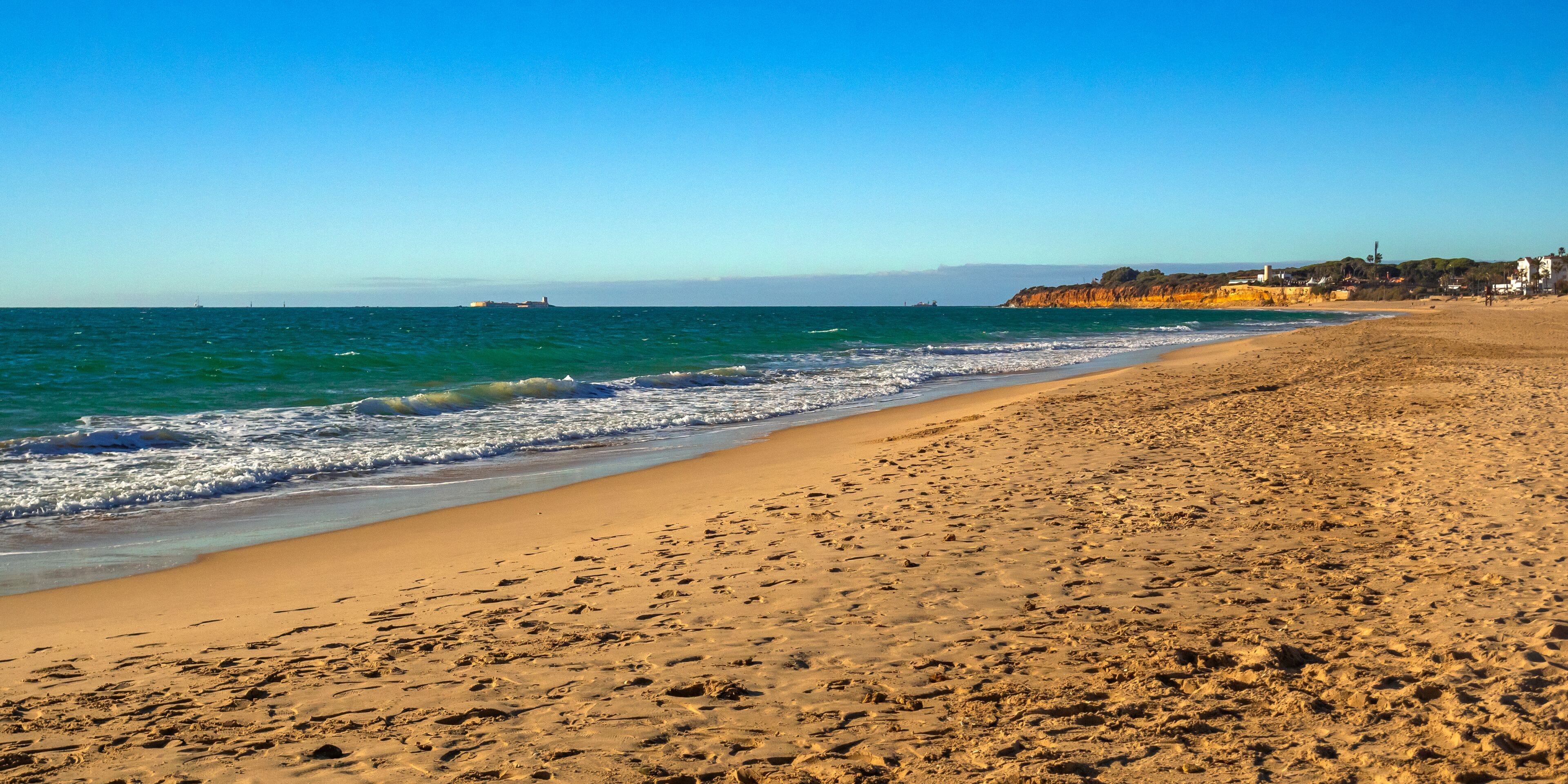 A sandy beach with waves, a clear blue sky, and buildings in the background.