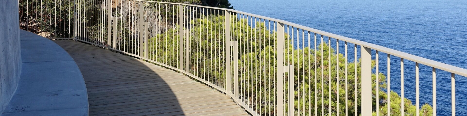 Un muelle de madera con barandillas blancas que conduce a una costa rocosa con abundante vegetación y el mar.