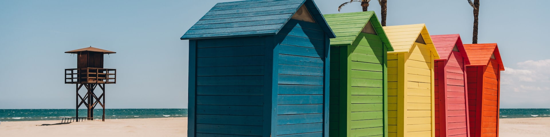 Una hilera de cabañas de playa coloridas en una playa arenosa, con palmeras y una torre de salvavidas al fondo.