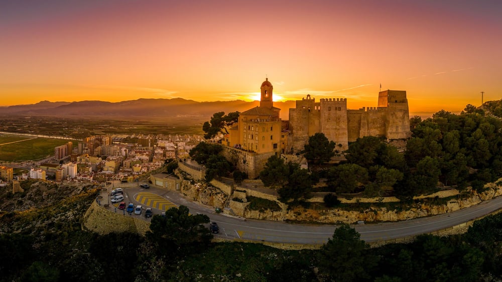 Eine historische Burg auf einem Hügel mit Blick auf eine Küstenstadt bei Sonnenuntergang.