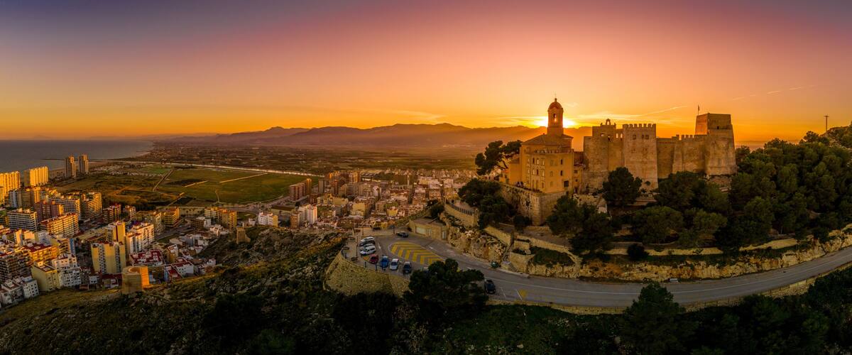 A historic castle on a hill overlooking a coastal city at sunset.