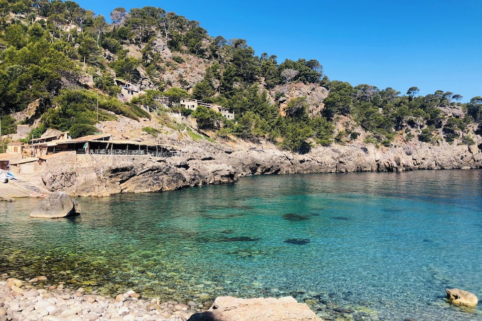 A coastal area with clear turquoise water, rocky shoreline, and buildings on a hillside.