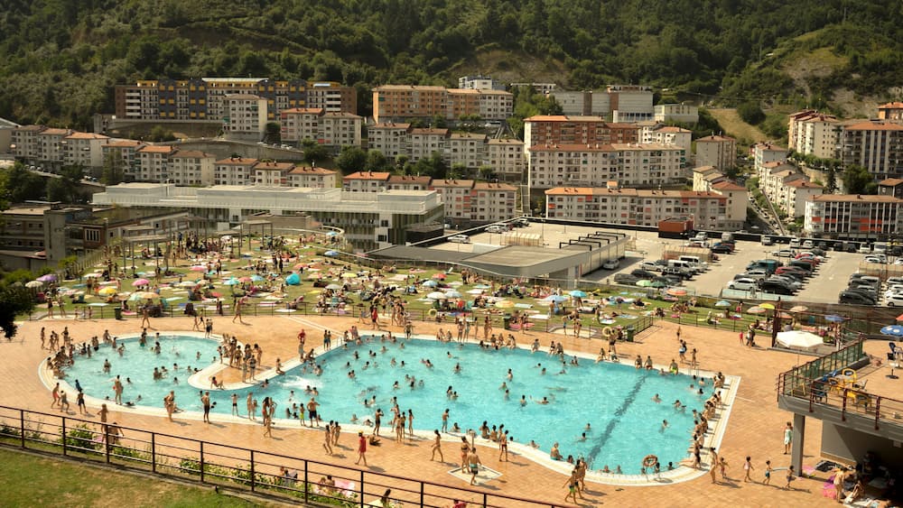 A large public swimming pool surrounded by a beach area with umbrellas and people.