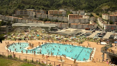 A large public swimming pool surrounded by a beach area with umbrellas and people.
