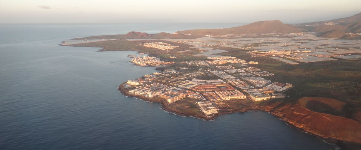 An aerial view of a coastal town with a dense cluster of buildings along the shoreline, surrounded by water and hilly terrain.
