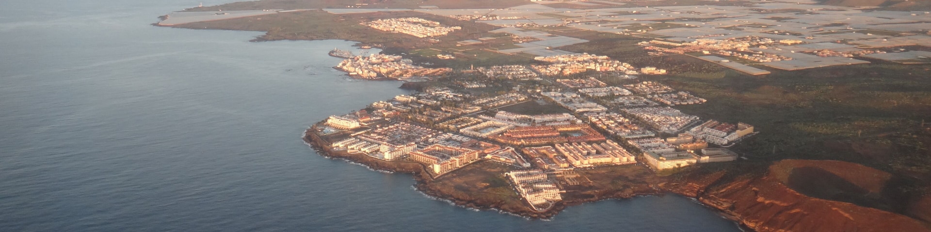 An aerial view of a coastal town with a dense cluster of buildings along the shoreline, surrounded by water and hilly terrain.