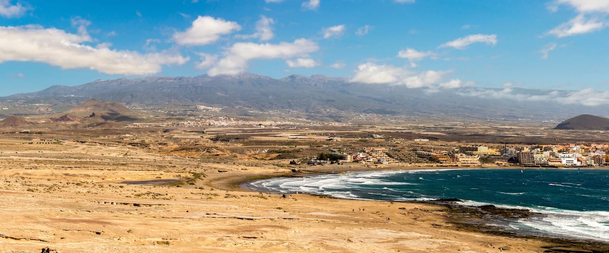 A coastal town with a beach, mountains in the background, and a clear sky.
