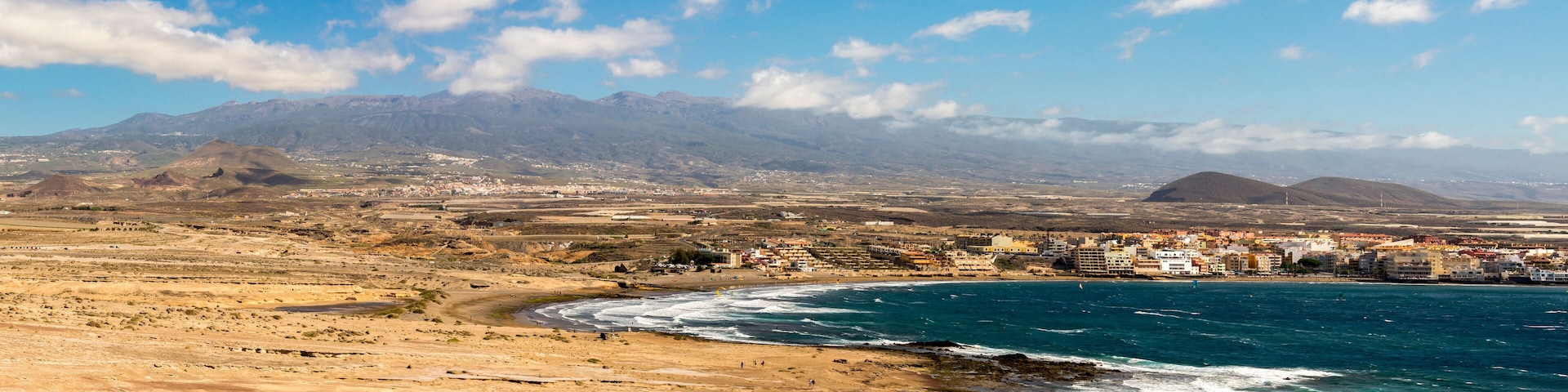 A coastal town with a beach, mountains in the background, and a clear sky.