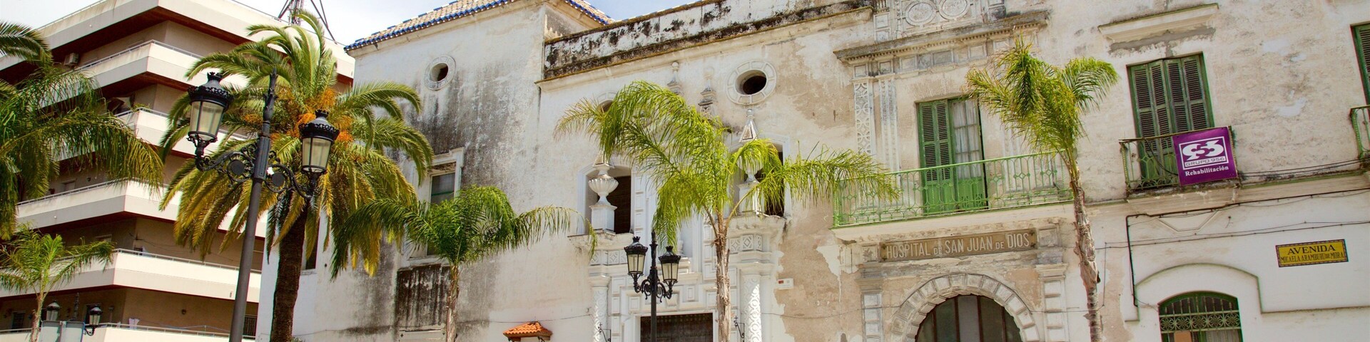 A historic building with a bell tower, palm trees, and a sign for 'Hospital de San Juan de Dios'.