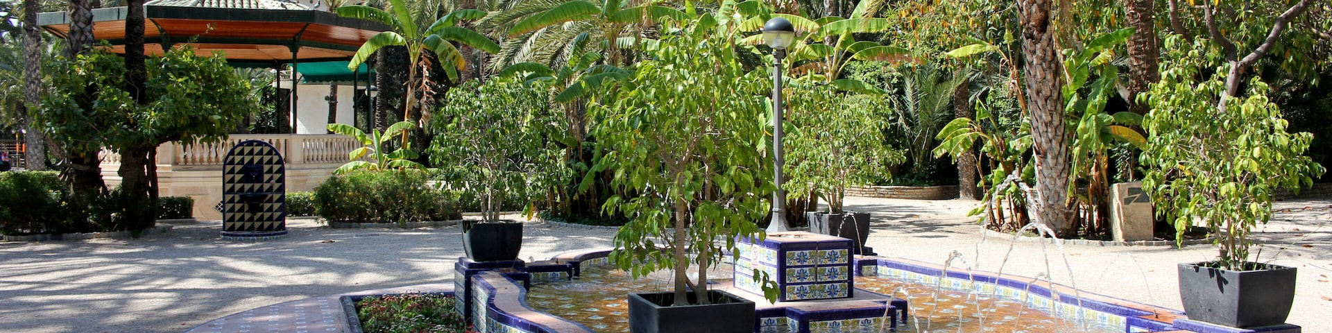 A fountain surrounded by tiled planters, palm trees, and a building with a tiled roof.