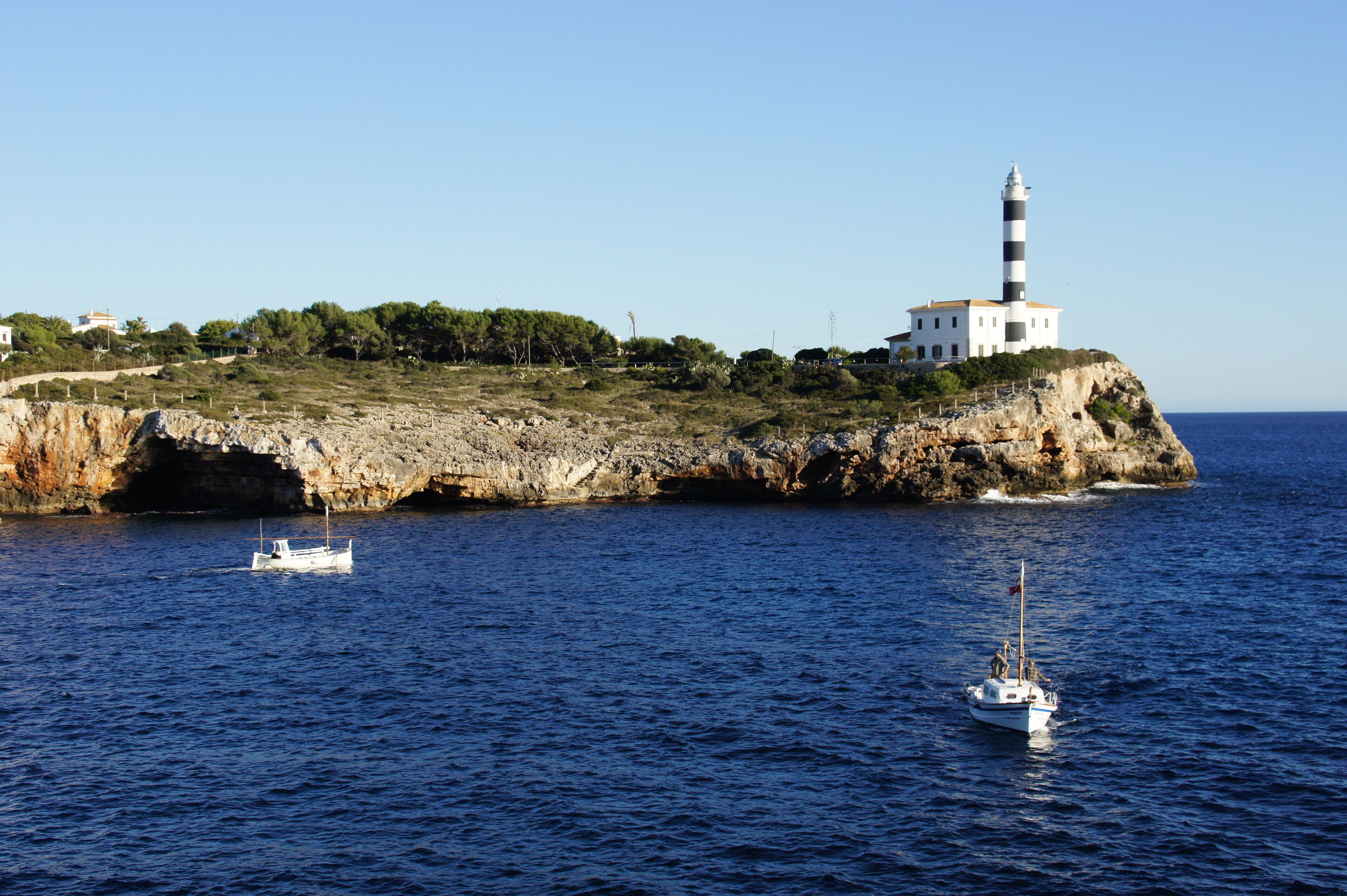 A coastal landscape with a lighthouse on a rocky outcrop, a small boat in the water, and a clear blue sky.