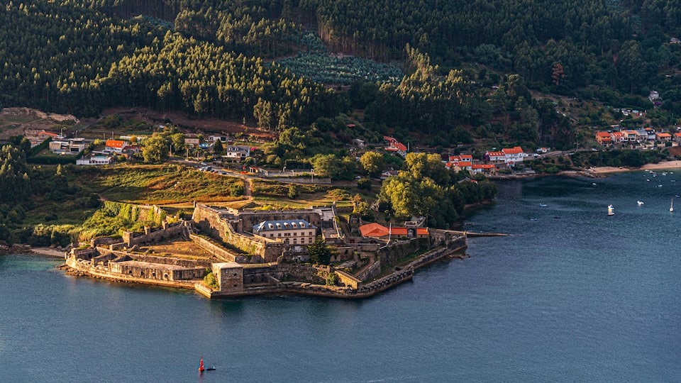 A coastal town with a fortified island, dense forested hills, and a calm sea.