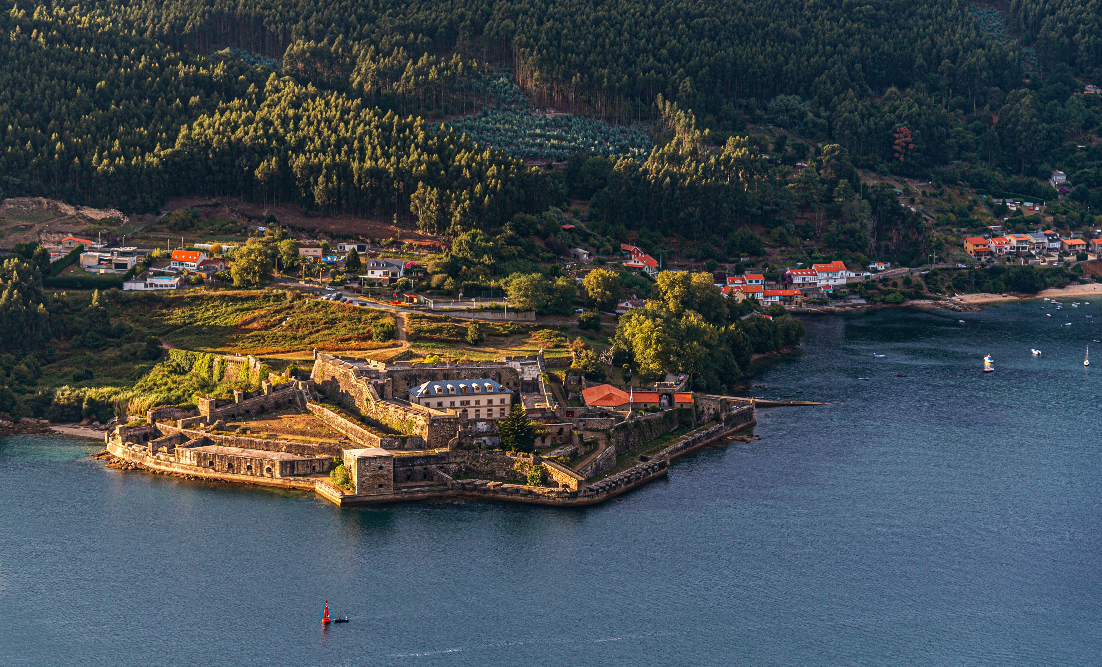 A coastal town with a fortified island, dense forested hills, and a calm sea.