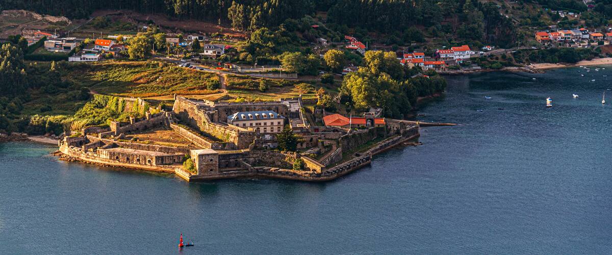 A coastal town with a fortified island, dense forested hills, and a calm sea.