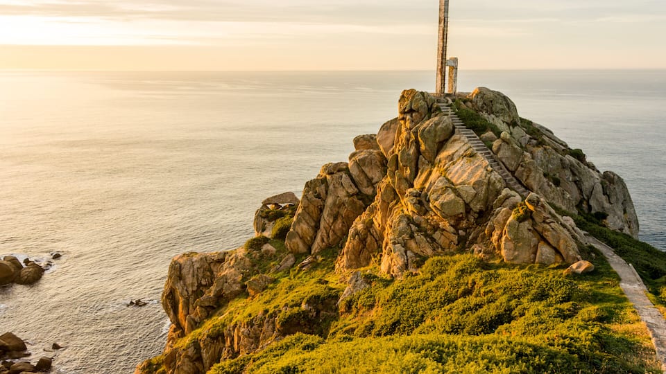 A coastal landscape with a prominent vertical structure on a rocky outcrop, a narrow path leading up to it, and the ocean in the background.