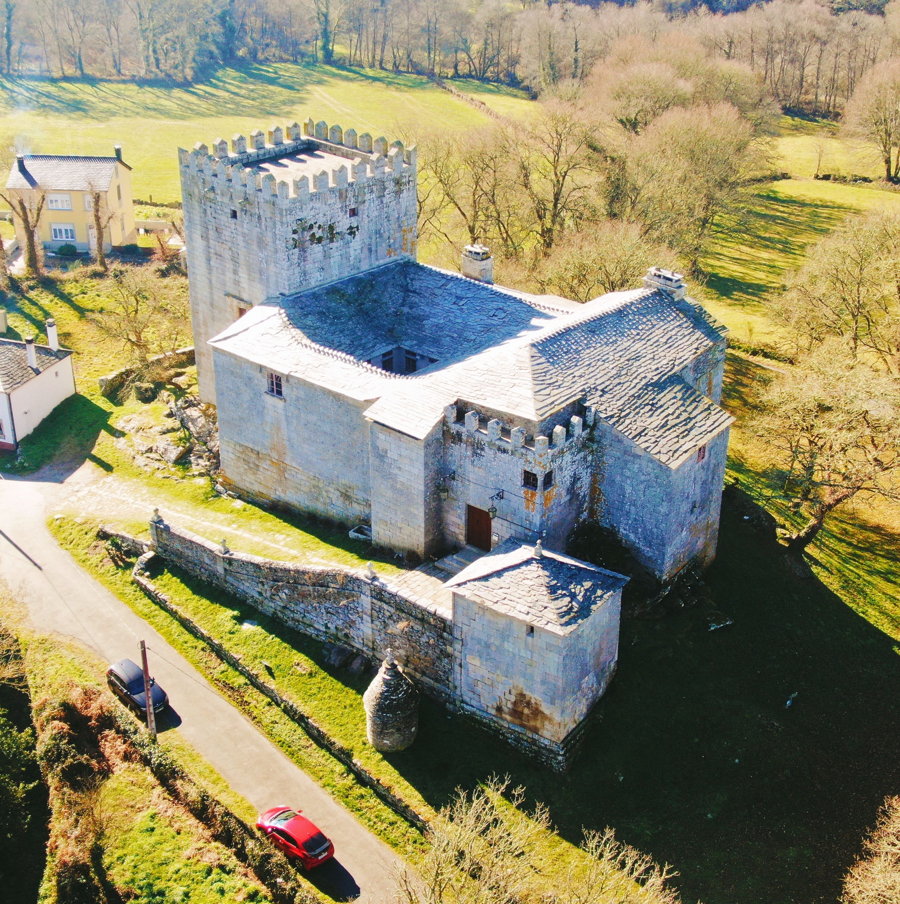 Un castillo medieval grande con un foso, rodeado por un paisaje rural con campos y árboles.