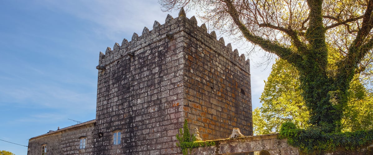 A historic stone tower with a battlement, a person with a red backpack standing in front, and a tree with green leaves to the right.