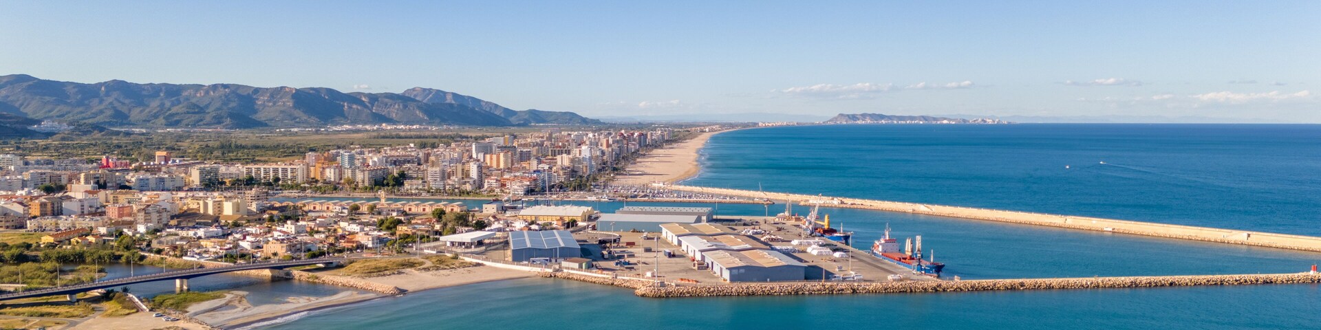 Un paisaje costero con playa, puerto y montañas al fondo.