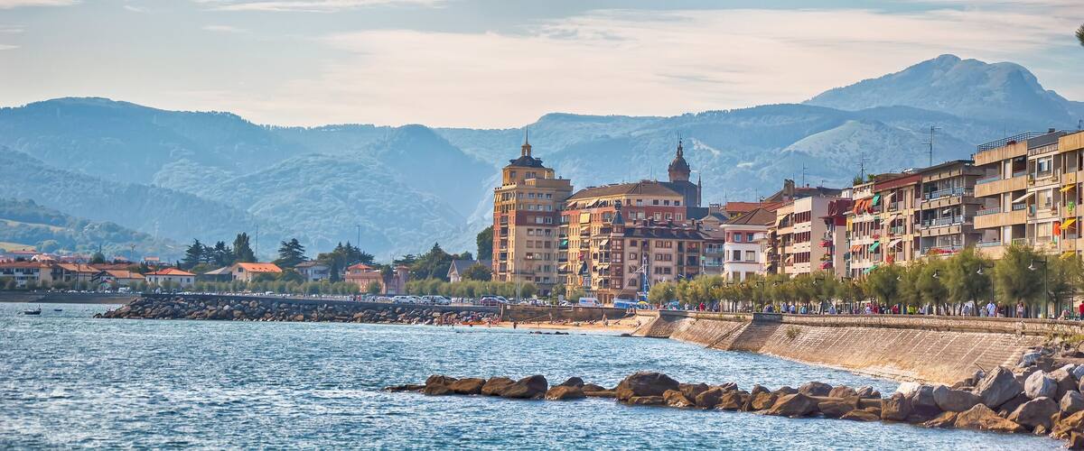 Un pueblo costero con un edificio destacado de torre, un litoral rocoso y montañas al fondo.