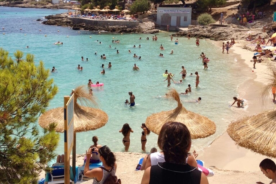 A crowded beach with clear turquoise water, straw umbrellas, and people swimming and sunbathing.