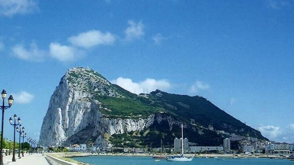 A coastal city with a prominent rocky mountain, a promenade, and boats in the water.