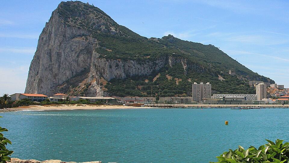 A coastal city with a prominent mountain, clear blue waters, and buildings along the shore.