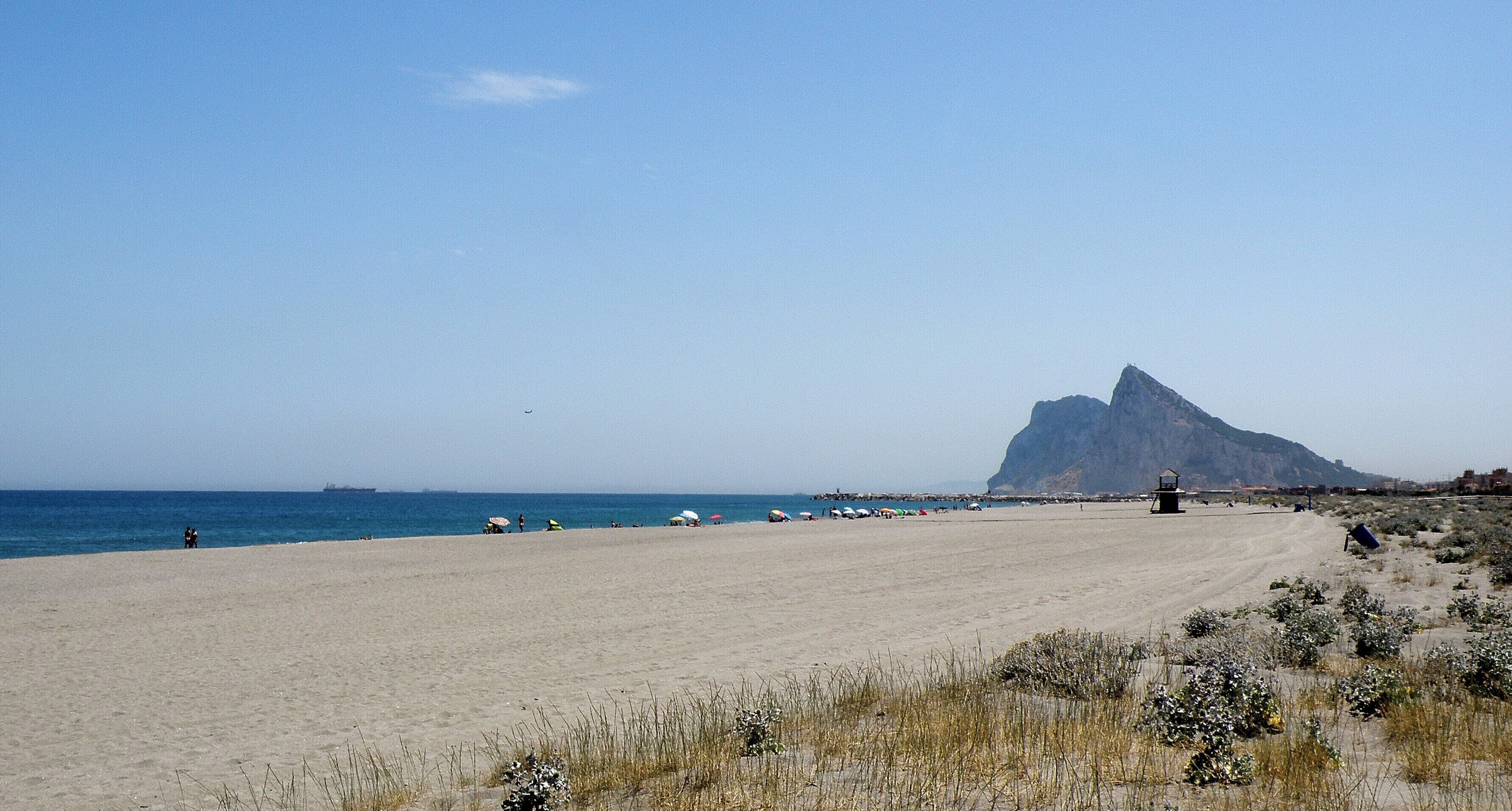 A beach with a large rock formation in the distance, a sandy shore, and a few people and umbrellas.