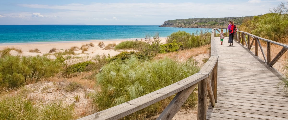 A wooden boardwalk leading to a beach with clear blue water and a distant hill.