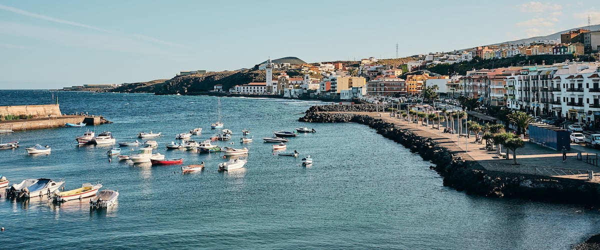 A coastal town with boats docked along the waterfront, a promenade, and buildings in the background.