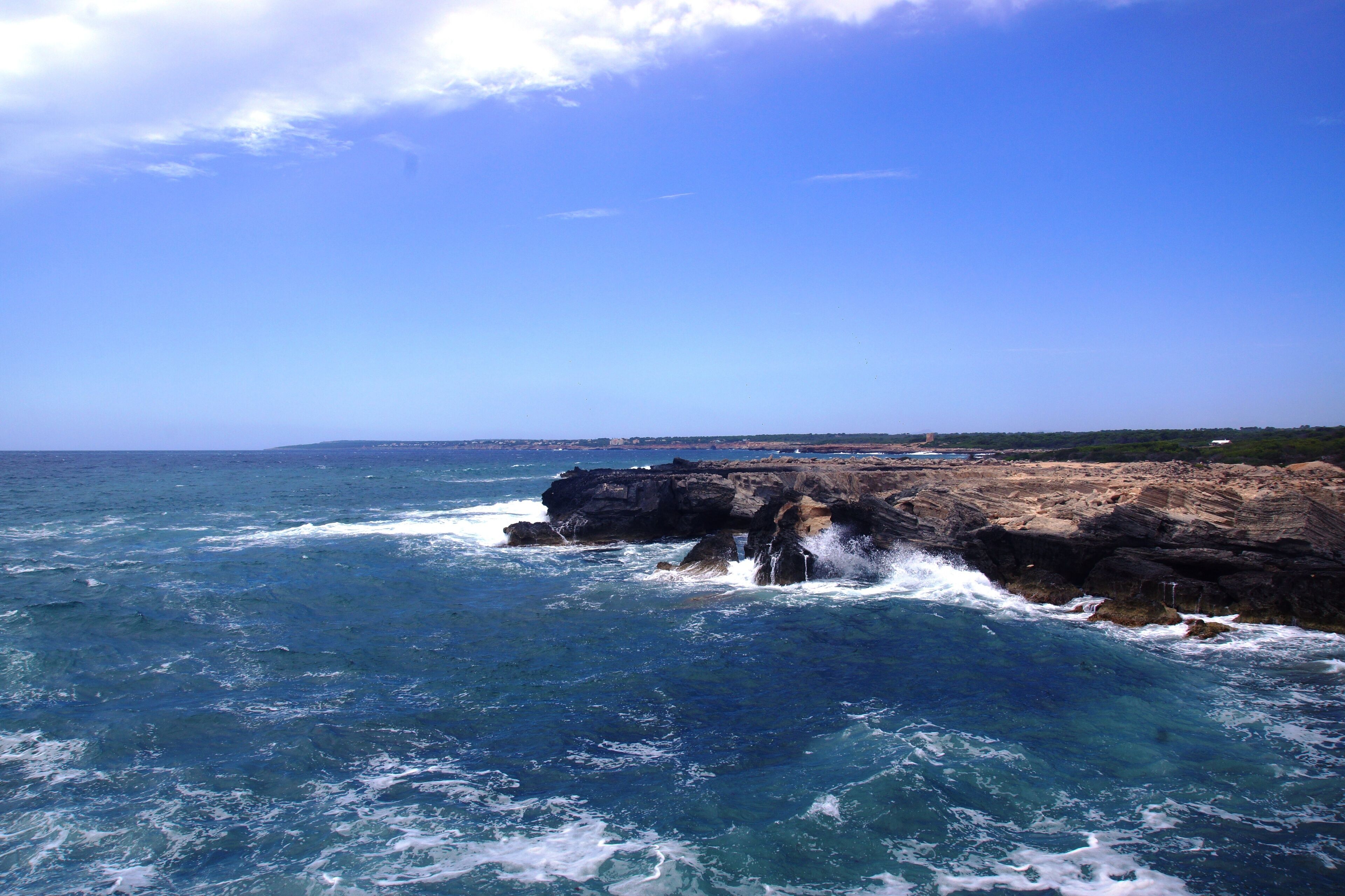 A rocky coastline with waves crashing against the rocks under a clear blue sky.