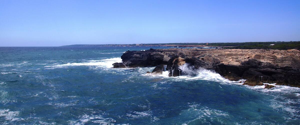 A rocky coastline with waves crashing against the rocks under a clear blue sky.