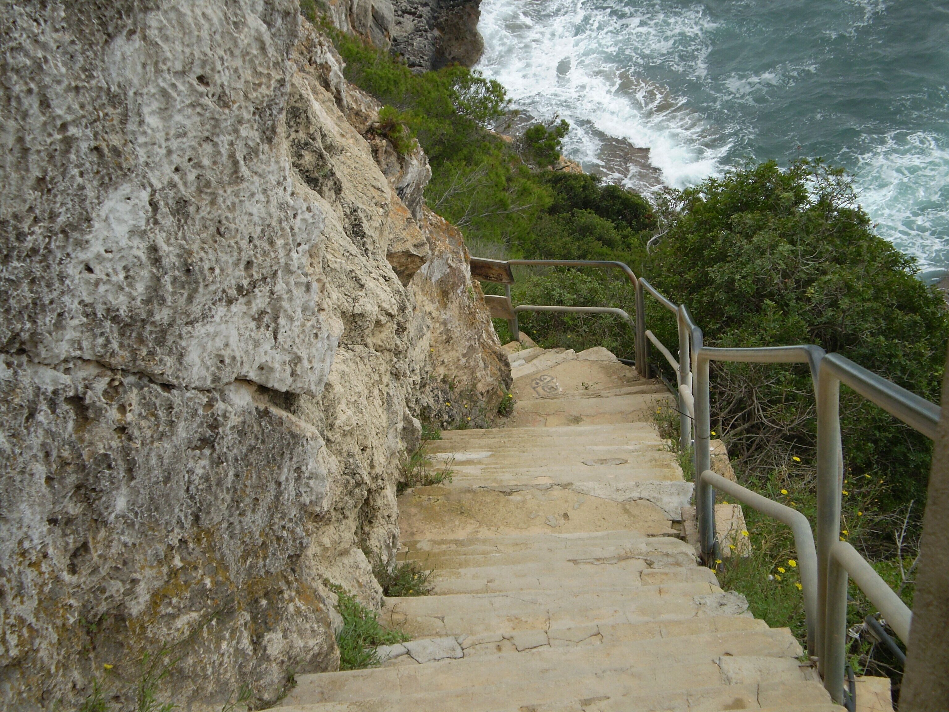 A stone staircase leading down a rocky cliffside with a railing, overlooking the ocean.