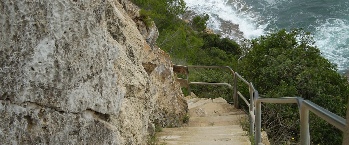 A stone staircase leading down a rocky cliffside with a railing, overlooking the ocean.
