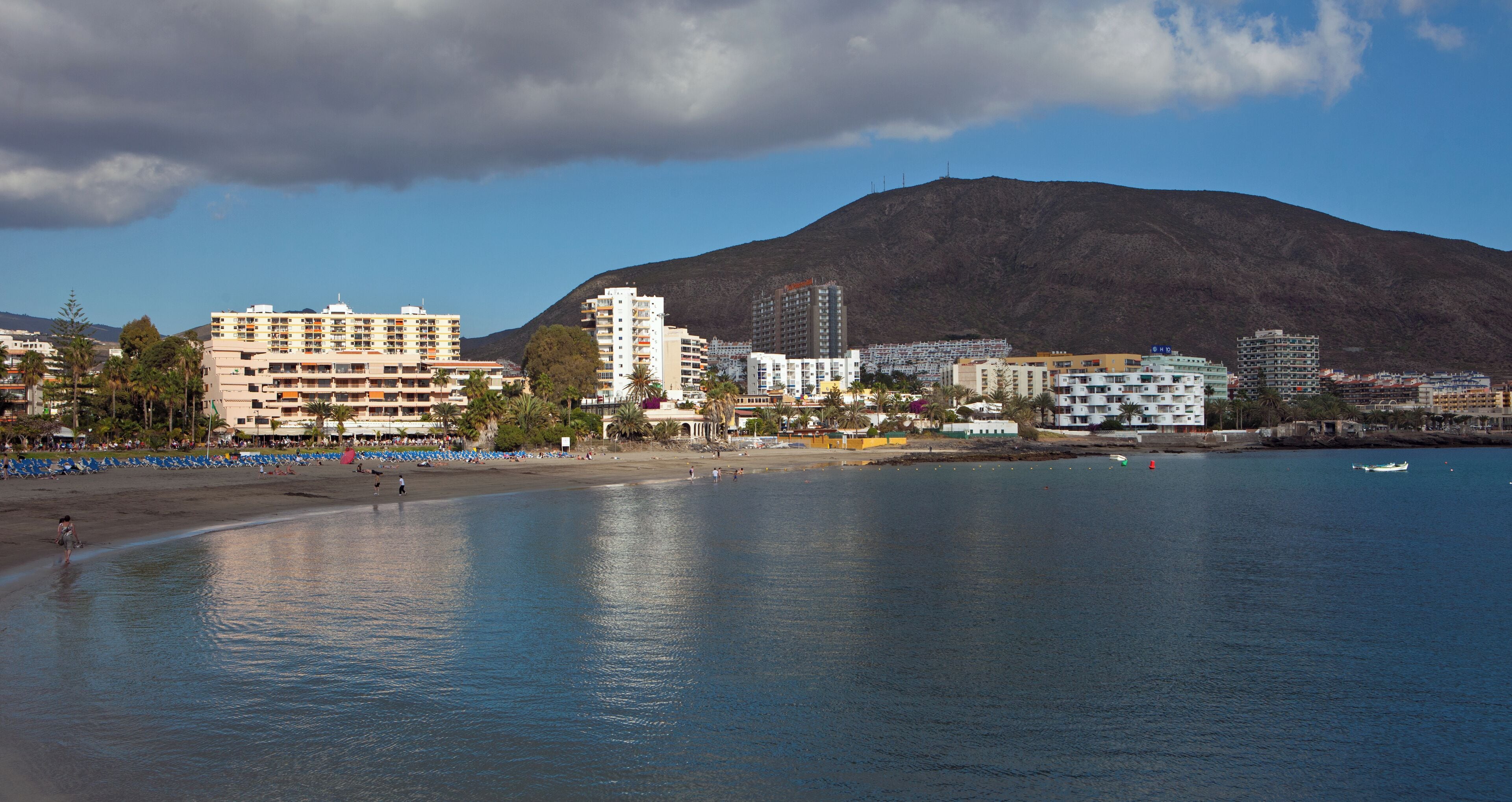 A coastal cityscape with a prominent mountain in the background, a sandy beach with umbrellas, and several multi-story buildings.