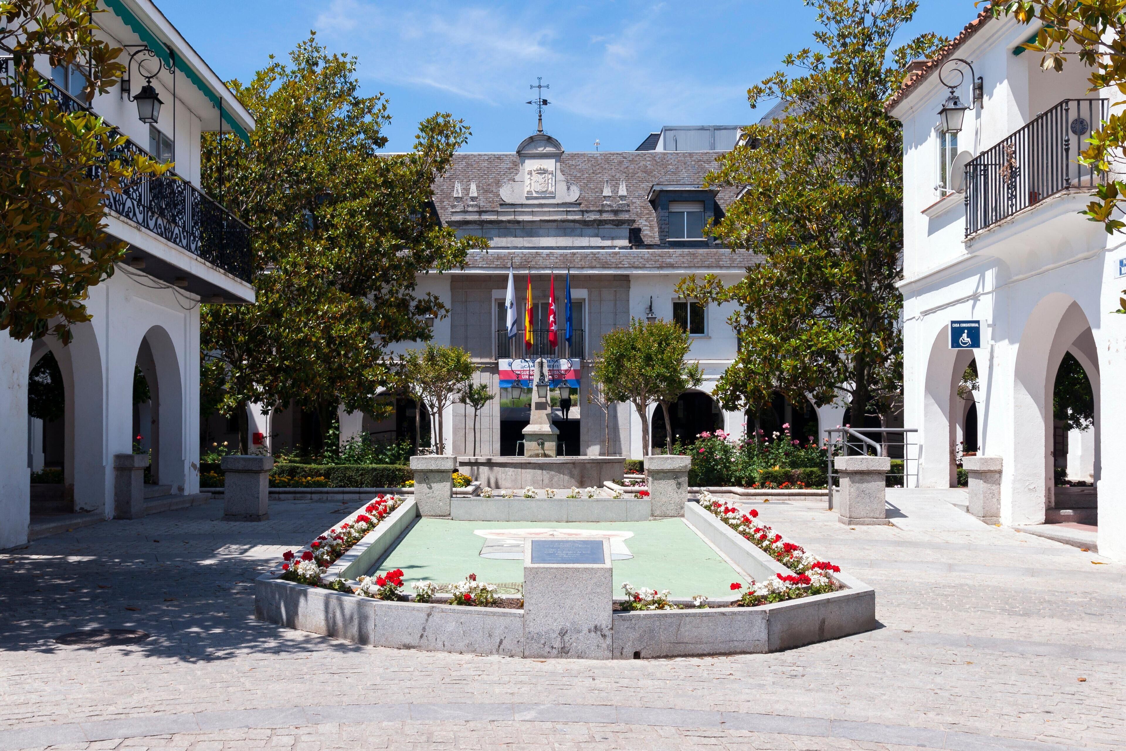 A plaza with a central fountain, surrounded by white buildings with arches and balconies.